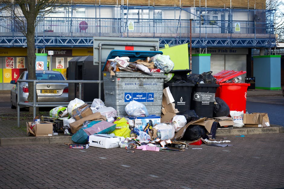 A large pile of mixed household waste and recyclables accumulated in front of a commercial building, featuring open and overflowing black, green, and red rubbish bins filled with paper, cardboard, pla